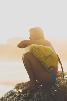 a woman sitting on top of a rock next to the ocean holding a surfboard