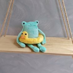 a stuffed frog sitting on top of a yellow duckling in front of a gray wall