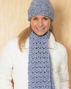 a woman wearing a blue knitted scarf and beanie smiles while standing in front of a wooden wall