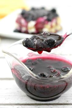 a spoon full of blueberry cheesecake sauce on top of a glass bowl with another dish in the background