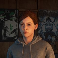 a woman in a grey hoodie standing next to posters on the wall and looking at the camera