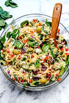 a glass bowl filled with pasta salad on top of a marble counter next to a wooden spoon