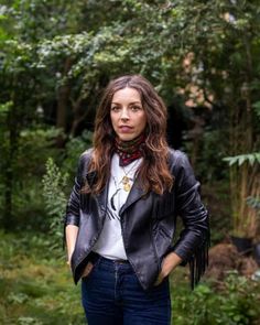 a woman standing in the woods wearing a black leather jacket and red scarf around her neck