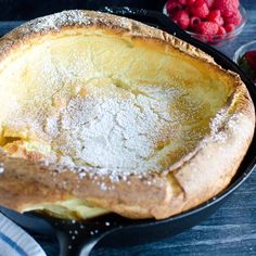 a pie in a cast iron skillet with powdered sugar on top and raspberries next to it