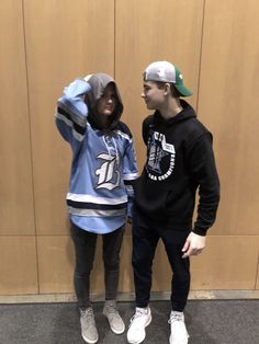 two young people standing next to each other in front of a wooden wall wearing hoodies and sweatshirts