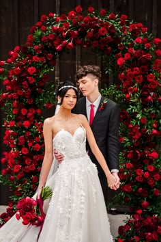 a bride and groom standing in front of a red rose arch with roses on it