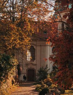 two people walking down a path in front of an old building with autumn leaves on the ground