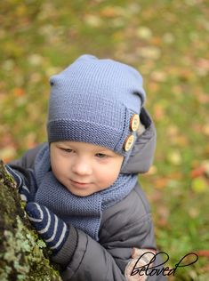 a young boy wearing a blue hat and scarf standing next to a tree trunk in the woods