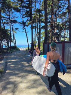 two women carrying surfboards down a road next to trees and the ocean in the background