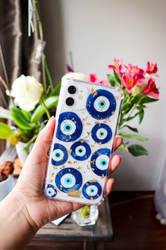 a hand holding an iphone case with blue and white eyeballs on it, in front of flowers