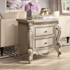 a white table with drawers and flowers on it in front of a couch next to a window