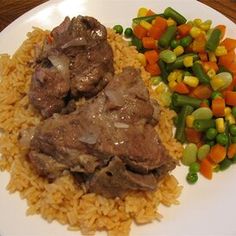 a white plate topped with meat and rice next to vegtables on a wooden table