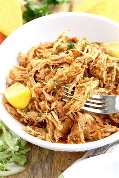 a white bowl filled with shredded chicken and lemon wedges on top of a wooden table