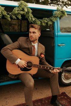 a man in a suit and tie sitting on the ground playing an acoustic guitar next to a blue van