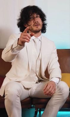 a man in a white suit sitting on a couch pointing his finger at the camera