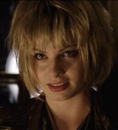 a woman with blonde hair wearing a leather jacket and looking at the camera in a dimly lit room