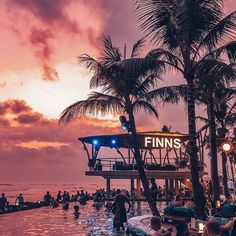 people are swimming in the water near palm trees and boats at sunset or sunrise time