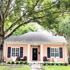 a pink house with black shutters on the front door and windows, sitting under a large tree