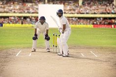 two men in white uniforms playing a game of cricket on a field with people watching from the stands