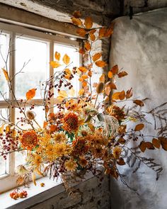 an arrangement of flowers in front of a window with leaves and branches on the windowsill