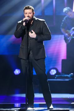 a male singer in a black suit and white shoes singing into a microphone on stage