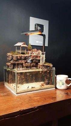 an aquarium with rocks and plants in it next to a coffee mug on a wooden table