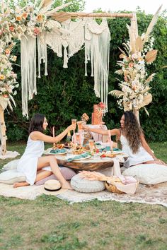 two women sitting at a table with food and drinks in front of an arch decorated with flowers