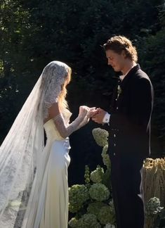 a bride and groom standing in front of some bushes looking at each other while exchanging their wedding rings