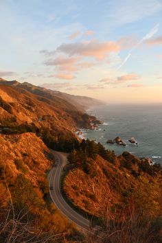 a winding road on the side of a mountain with ocean in the background at sunset