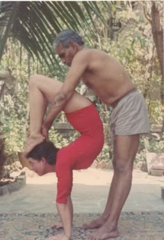 two men doing yoga poses in front of some trees and bushes, one man is bending over the other