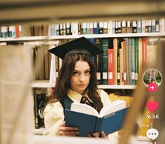 a woman in graduation cap and gown reading a book with bookshelves behind her