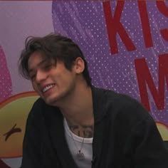 a young man sitting in front of a purple and red sign with the words kiss me on it