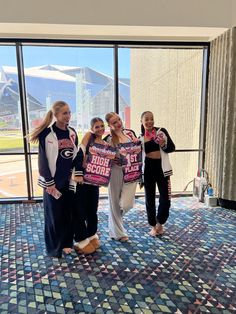 four girls standing in front of a window with their shirts on and one girl holding up a cell phone