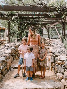 a woman standing with two children in front of a stone wall and pergolated area