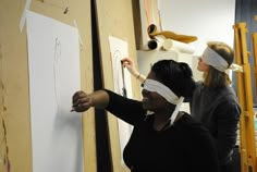 two women in blindfolds are drawing on a wall with white paper and scissors