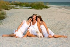 three women in white dresses sitting on the beach with a dog and smiling at the camera