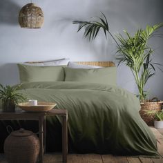 a bed with green sheets and pillows next to a potted plant on a table