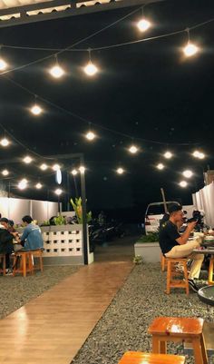 people sitting at tables under lights in an open area with wooden benches and stools