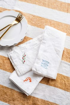 three white napkins with embroidered designs on them next to a plate and gold fork