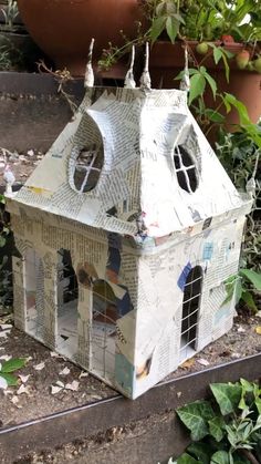 a house made out of newspaper sitting on the ground next to some plants and potted plants