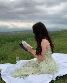 a woman sitting on top of a white blanket reading a book in a green field