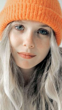 a woman with long white hair wearing an orange knitted hat and looking at the camera