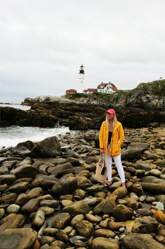 a woman in yellow jacket standing on rocks next to the ocean with lighthouse in background