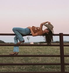 a woman leaning on a fence with her legs crossed and wearing cowboy boots, jeans and a brown shirt