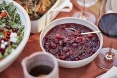 a table topped with plates and bowls of food next to glasses of red wine on top of a wooden table