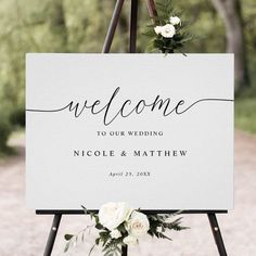 a welcome sign with flowers and greenery hanging from it's easel in front of a gravel road