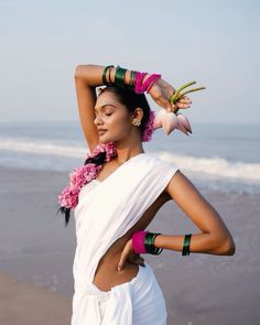 a woman with flowers in her hair standing on the beach next to the ocean wearing white clothing and colorful bracelets