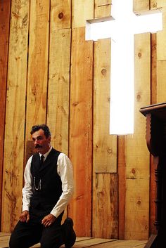 a man sitting on the floor in front of a wooden wall with a cross above it