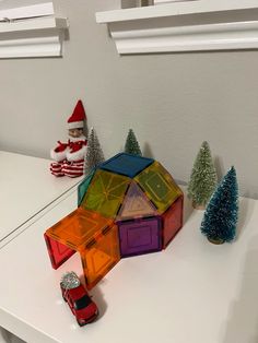 a toy house made out of colored glass blocks on a white table next to christmas trees