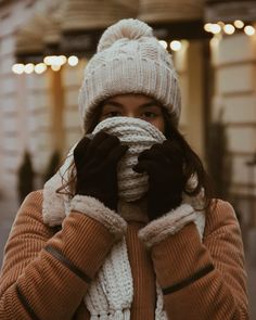 a woman covers her face with gloves and mittens while standing in front of a building